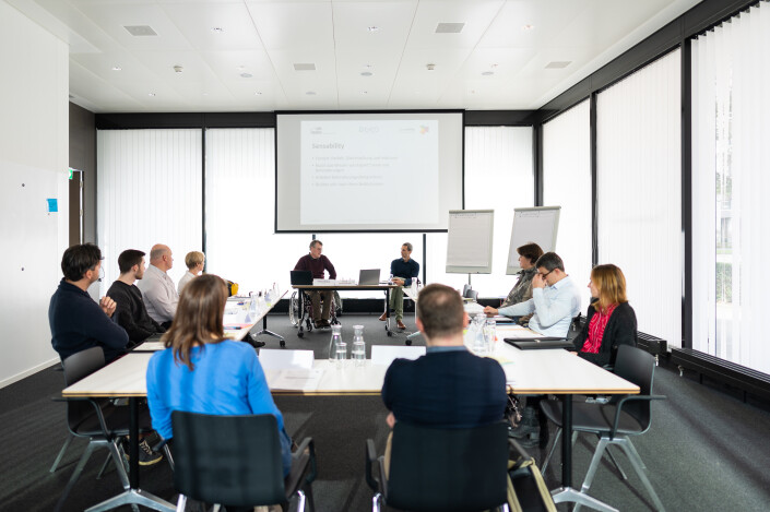 Siituation in einem Workshopraum mit Teilnehmenden, die in Hufeisenbestuhlung an Tischen sitzen.