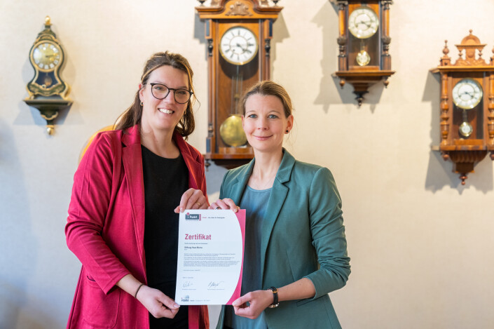 Zwei Frauen stehen vor einer Wand mit vielen Uhren und sie halten das iPunkt Zertifikat in die Kamera.