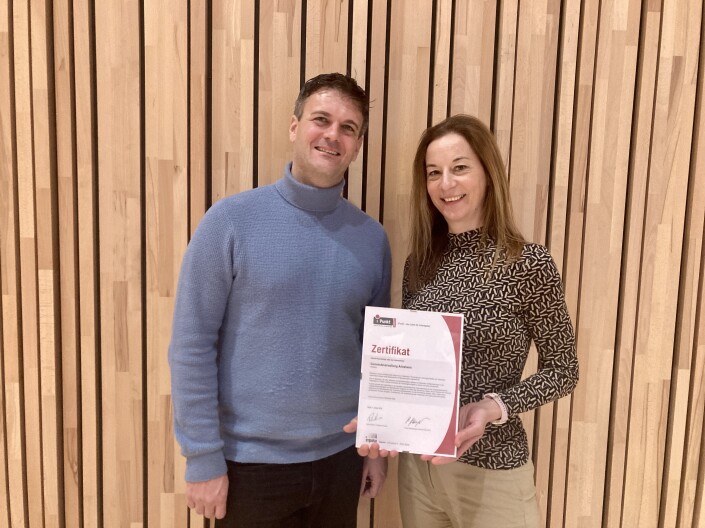 Claude Burgener (Mitarbeiter Stabsdienste/Projekte) und Tabea Studer (Leiterin HR und iPunkt-Beauftragte) stehen vor einer Holzwand und halten das iPunkt-Zertifikat in die Kamera.