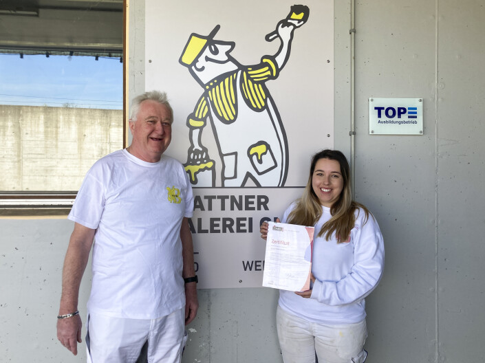 Thomas Blattner und Samira Forster stehen vor ihrem Logo und halten das Zertifikat in die Kamera.